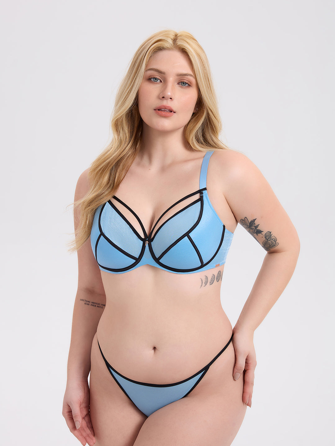 A blonde model wearing a light blue strappy lingerie set with black trim, consisting of a structured underwire bra and matching bikini bottoms, against a solid light gray background.