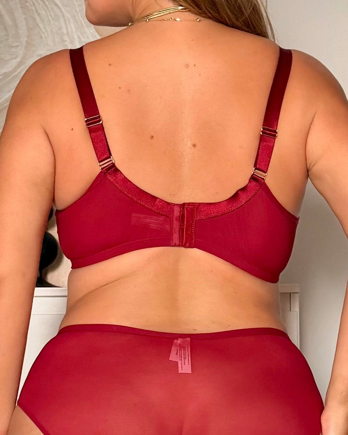 A woman shown from the back wearing a matching red lingerie set with a bra and high-waisted briefs, photographed indoors against a light wall and white cabinet.