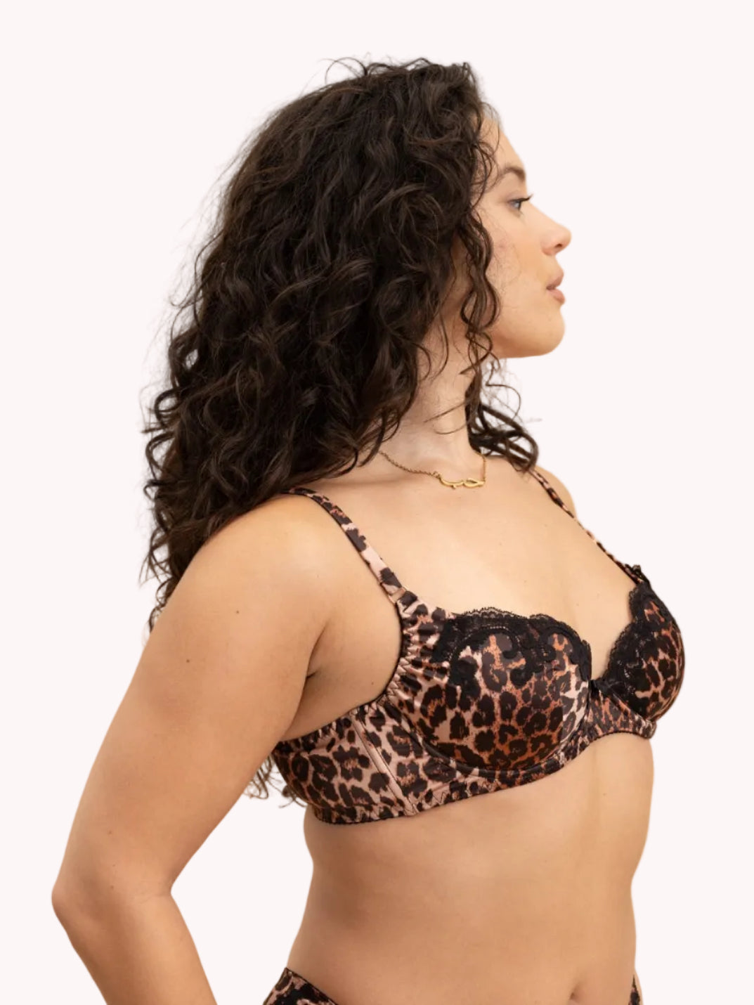 A woman modeling a leopard print lingerie set against a solid beige background. The set features a black and brown leopard print bralette with lace trim and matching low-rise bikini bottoms.