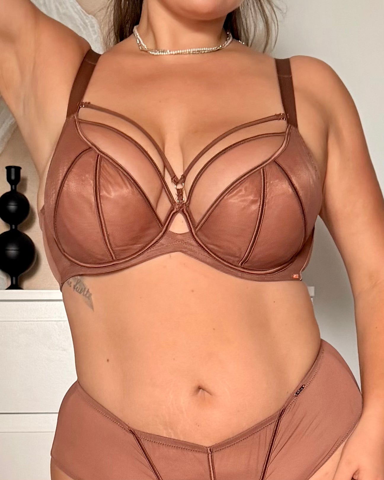 A woman wearing a brown strappy bralette and matching underwear, cropped from the neck to the hips against an indoor wall background.