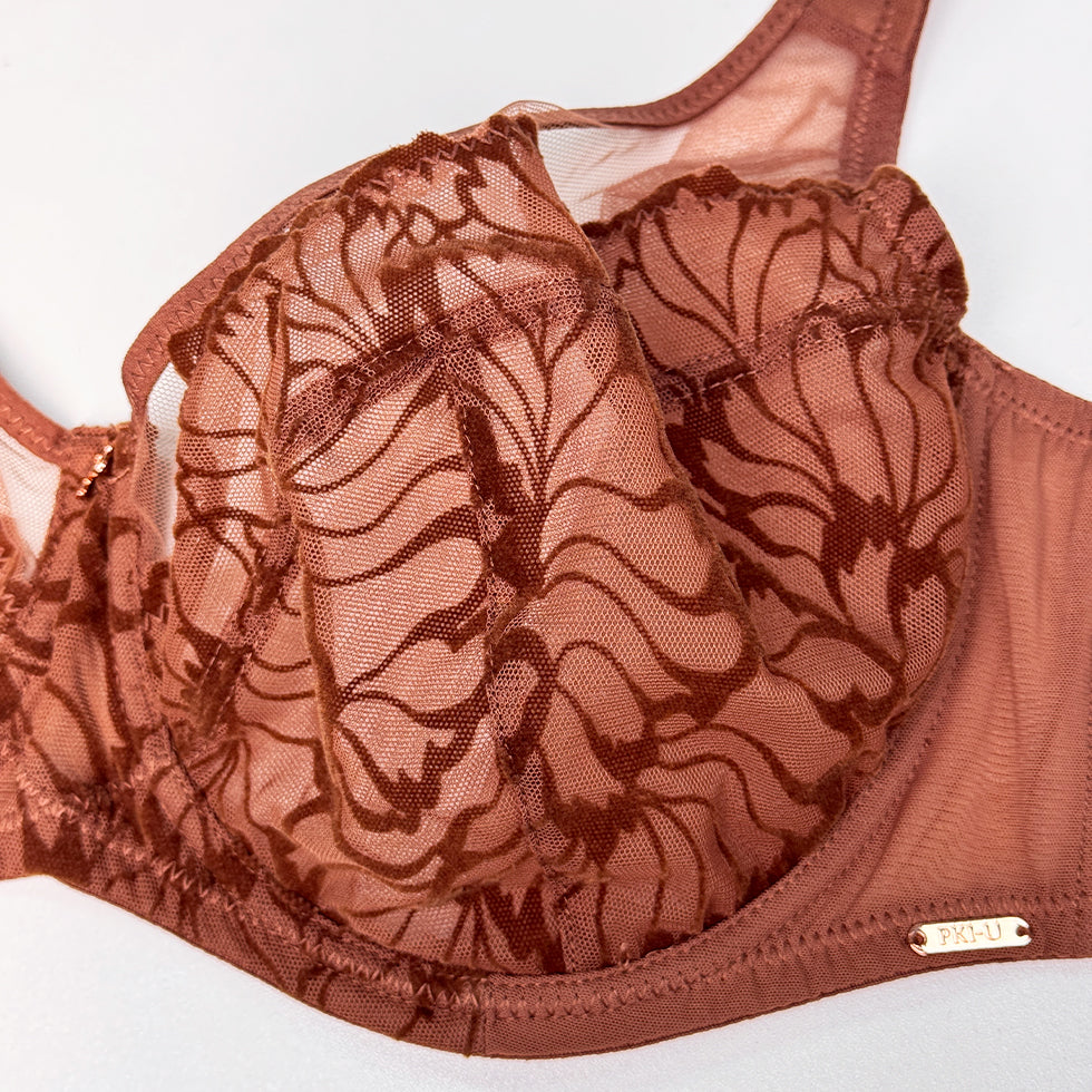 A close-up of a brown lace underwire bra with a floral pattern and a small gold-tone PROAUL logo plaque on the underband, set against a plain white background.