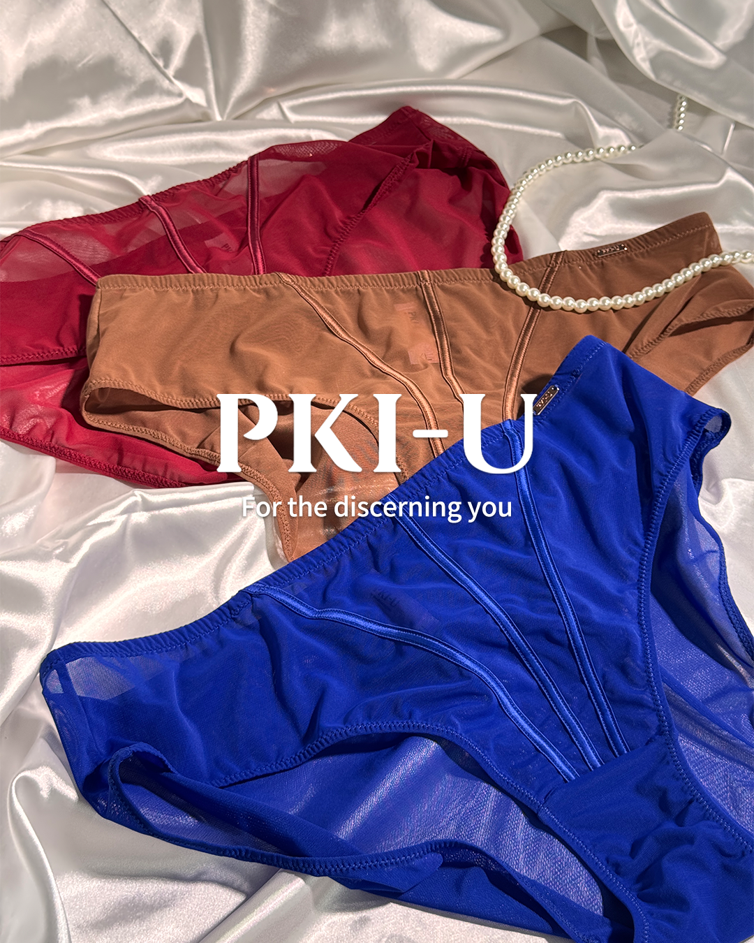 Three pairs of sheer mesh underwear in red, tan, and royal blue, arranged on a shiny silver satin background with a pearl necklace prop. White text overlay reads 'PKI-U For the discerning you'.