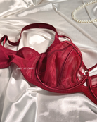 A deep red mesh and satin underwire bra laid flat on a white satin fabric background, with a pearl necklace partially visible in the top right corner.