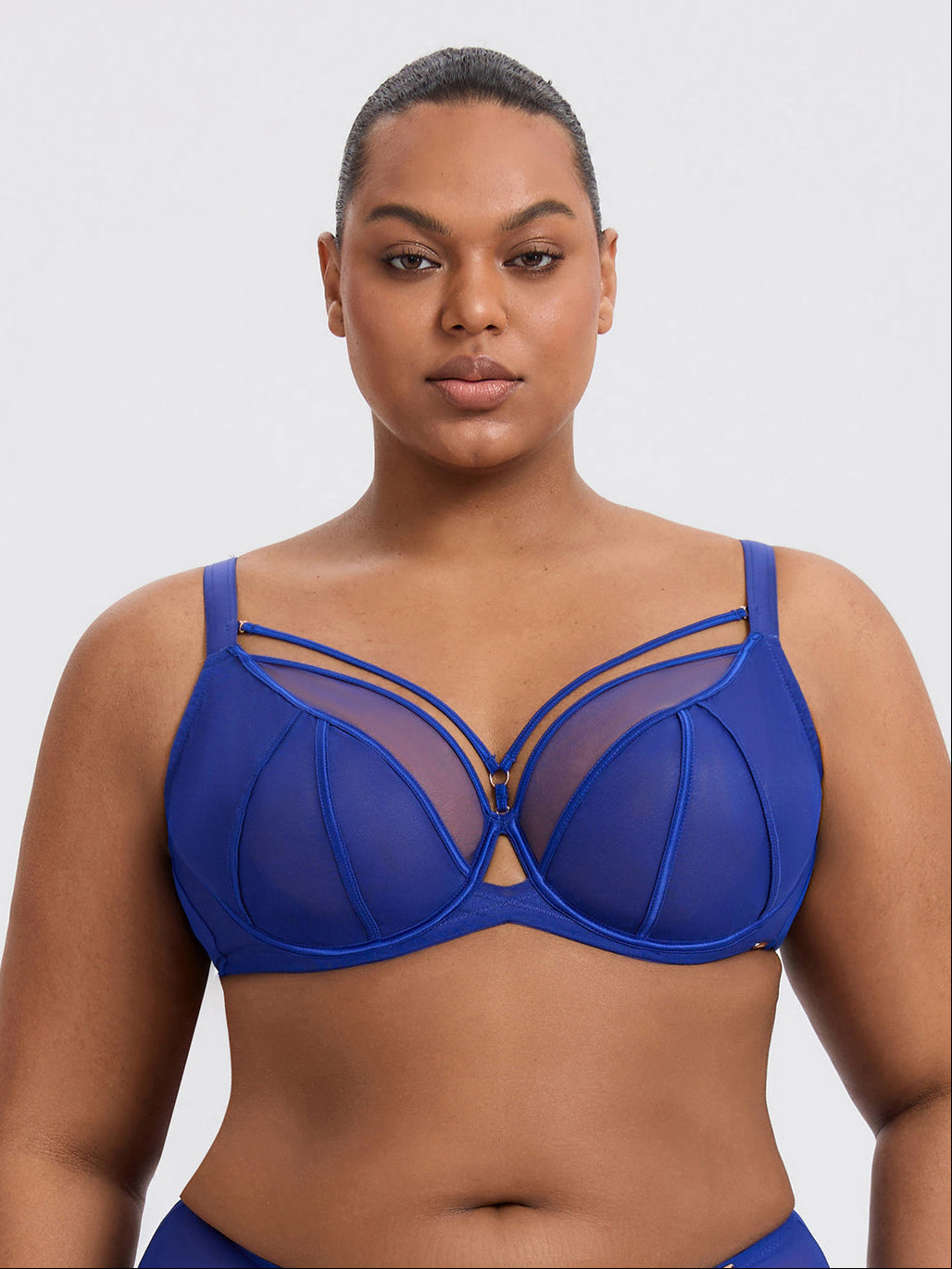 A plus-size model wearing a sapphire blue strappy mesh bra against a light gray studio background.