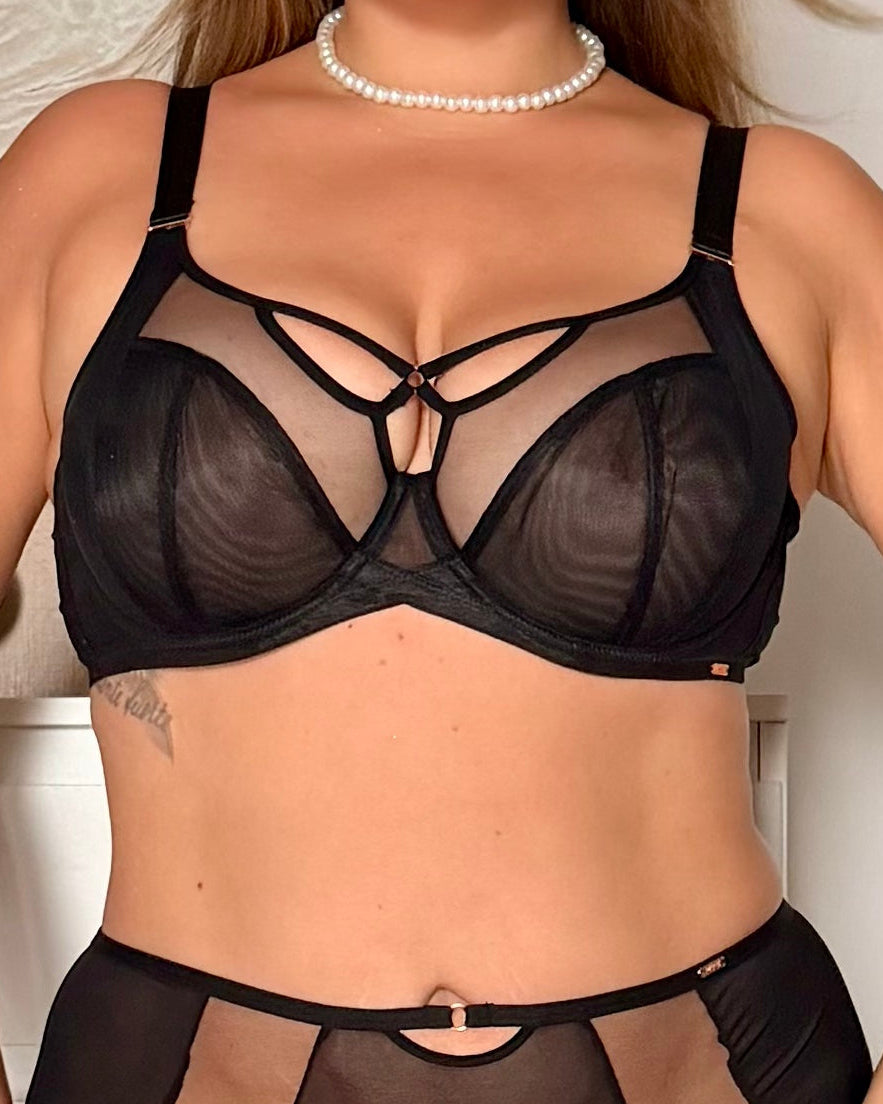 A woman modeling a black sheer mesh lingerie set with strappy details, featuring a bra and matching high-waisted briefs, against a light indoor background.