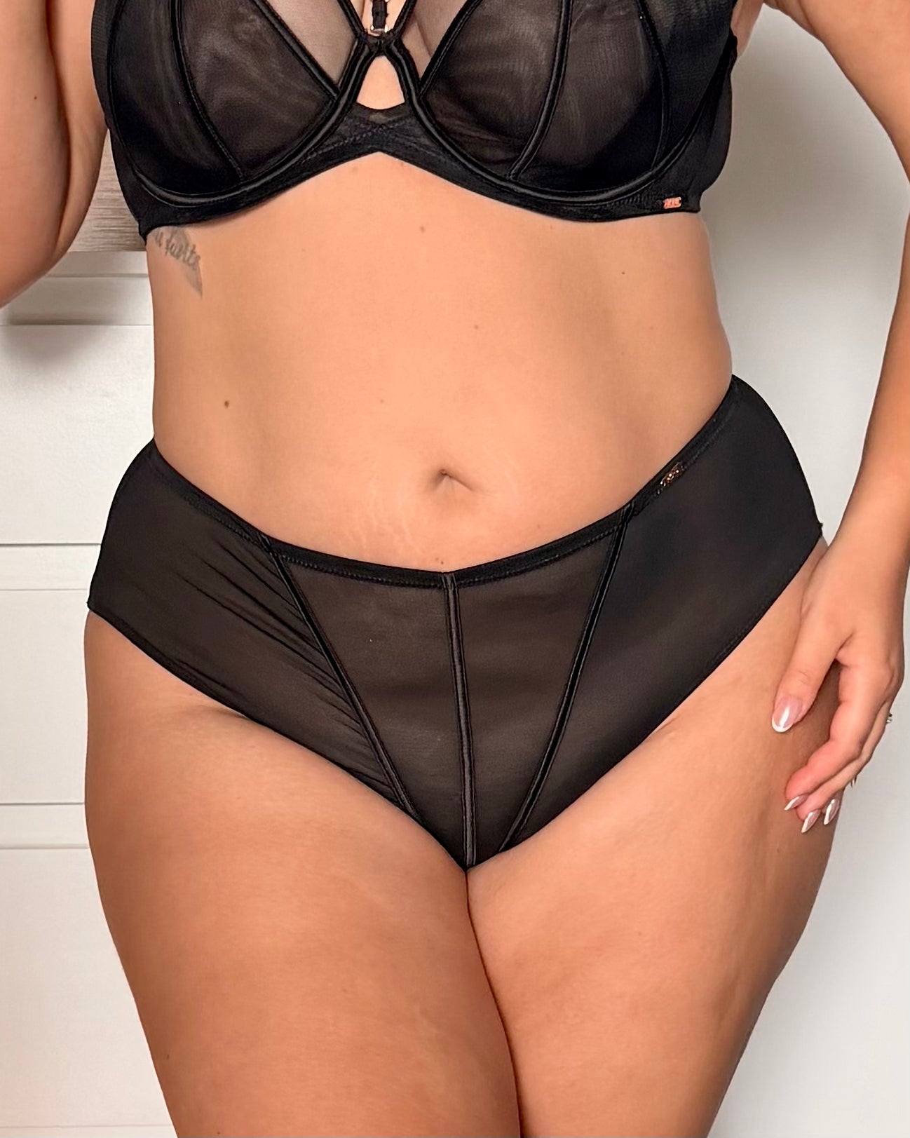 A close-up of a model wearing a black sheer mesh lingerie set with a strappy bra and matching high-waisted briefs against a white paneled wall.