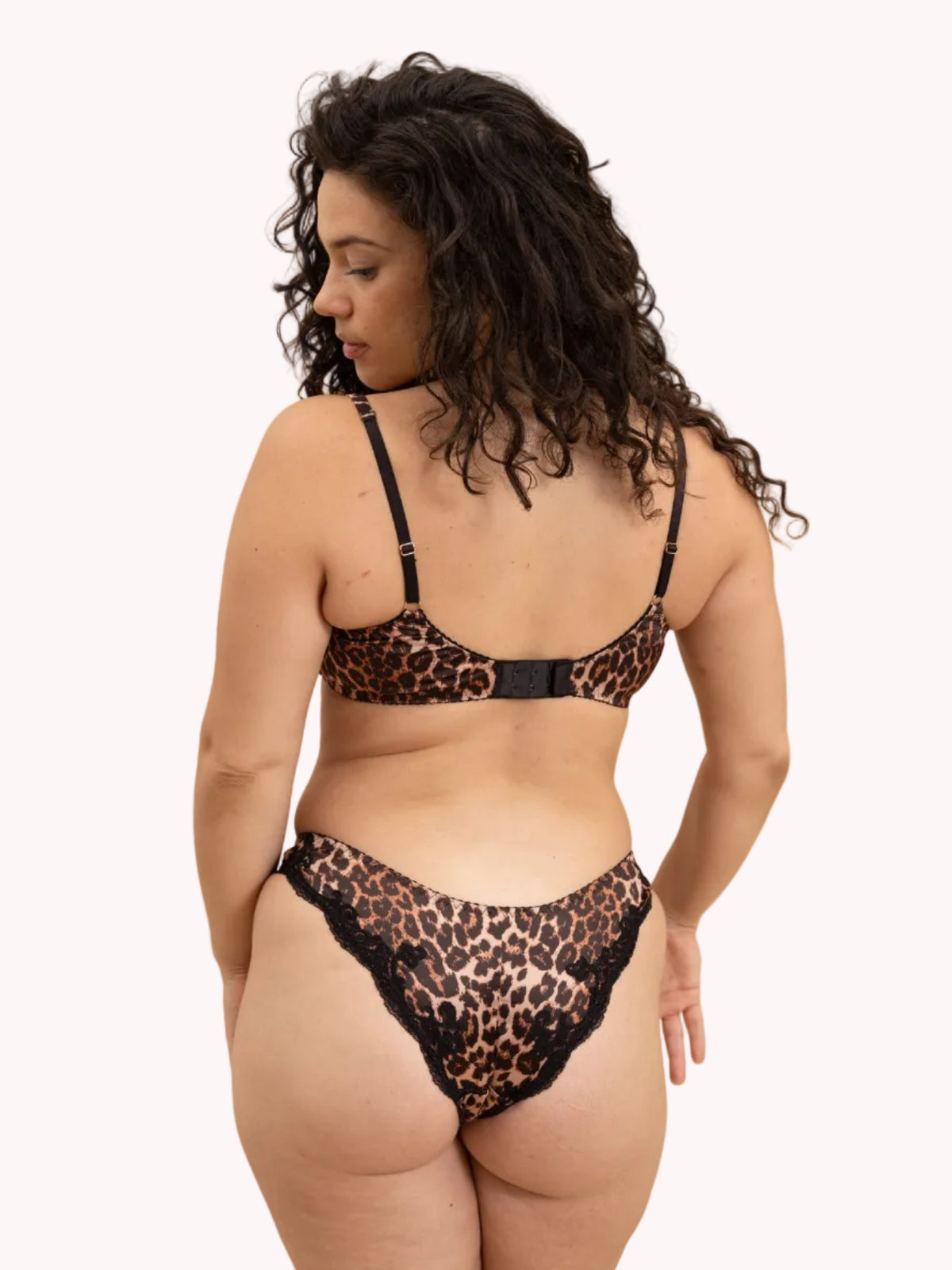 A woman with long, curly dark hair is shown from the back wearing a leopard print lingerie set with black trim, against a solid studio background.