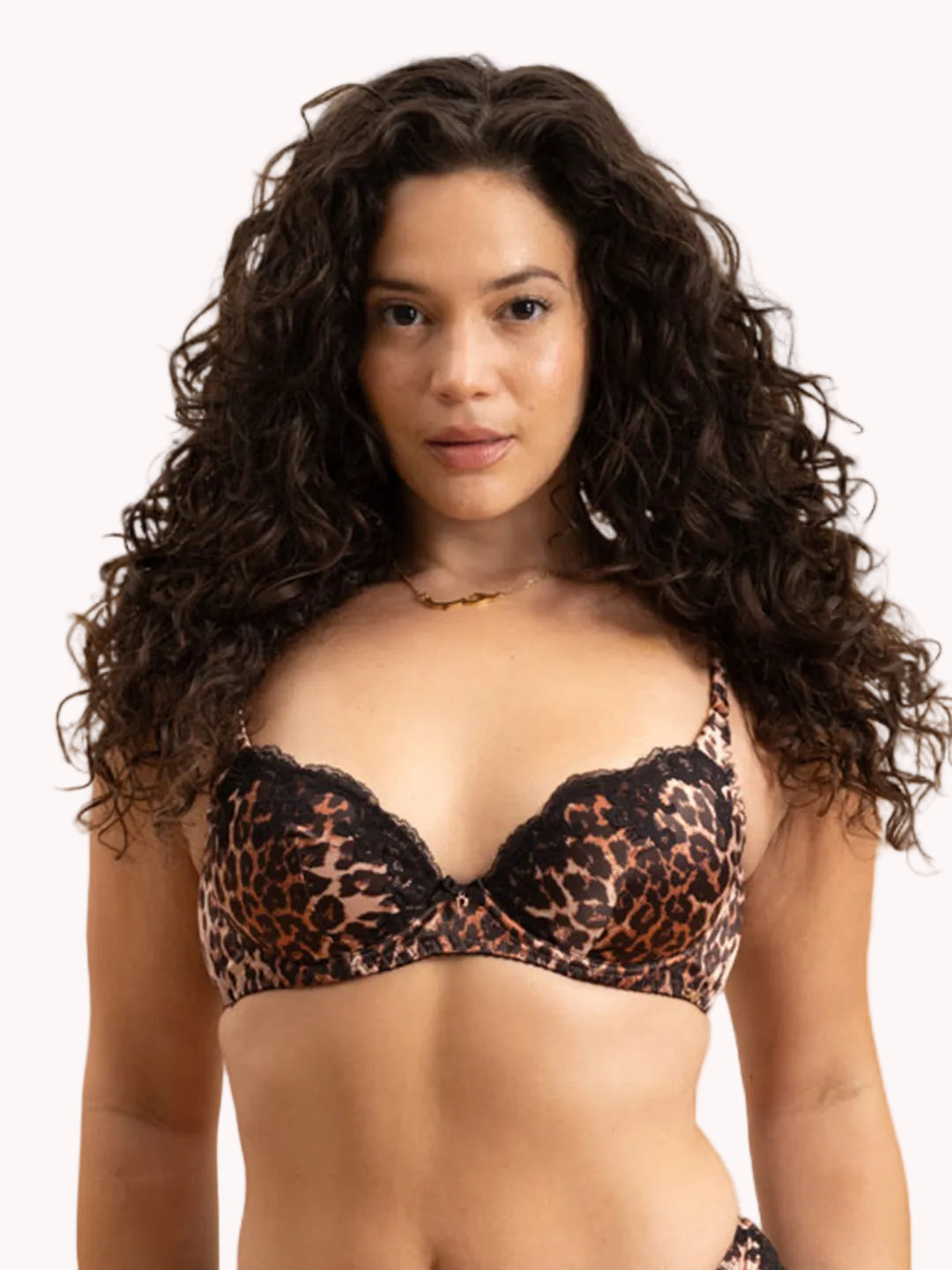 A woman with long, curly dark brown hair wearing a leopard print bralette with black lace trim, photographed against a solid white background.