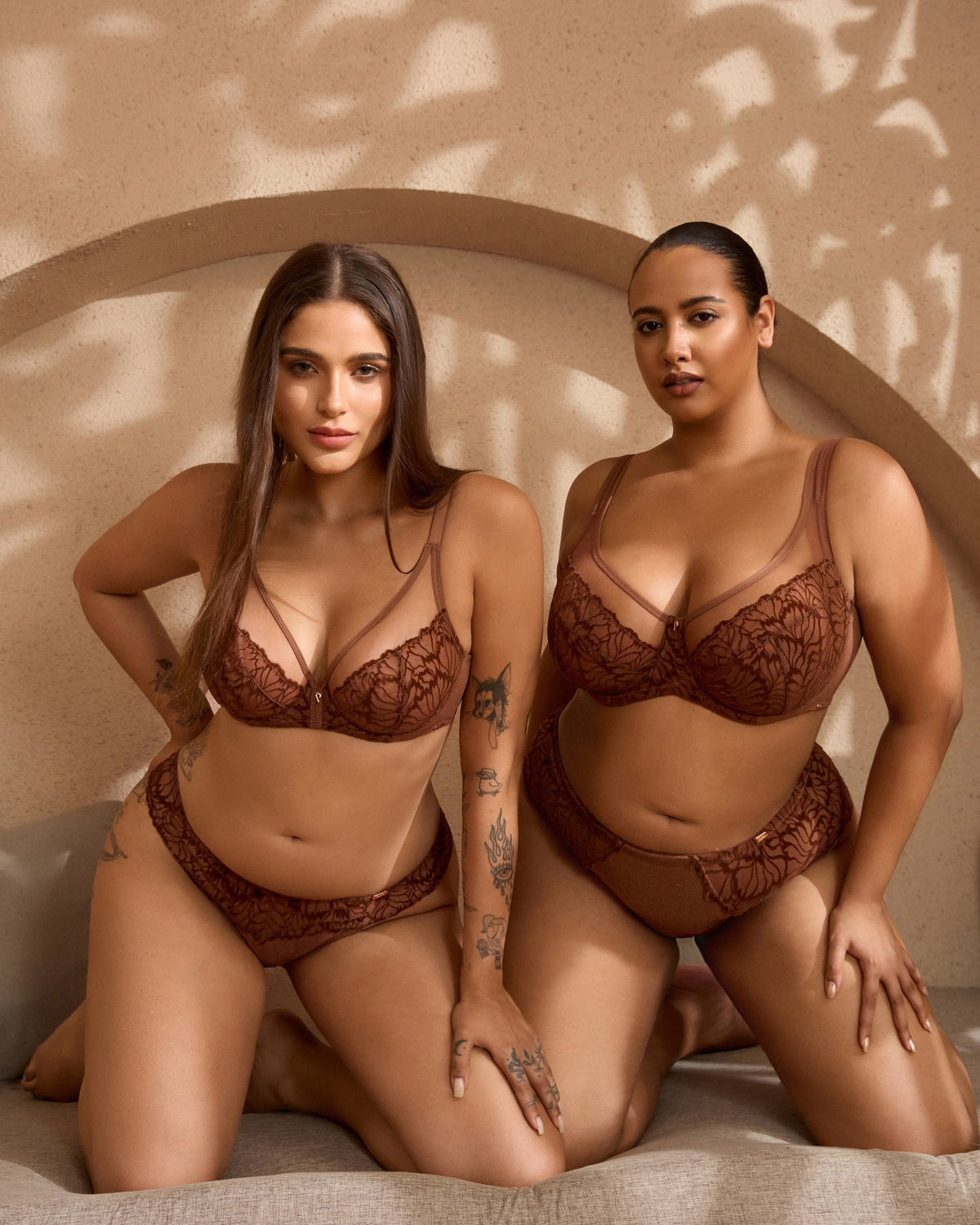 Two women modeling matching brown lace lingerie sets in a warm-toned studio setting with a textured beige backdrop and leaf shadows.