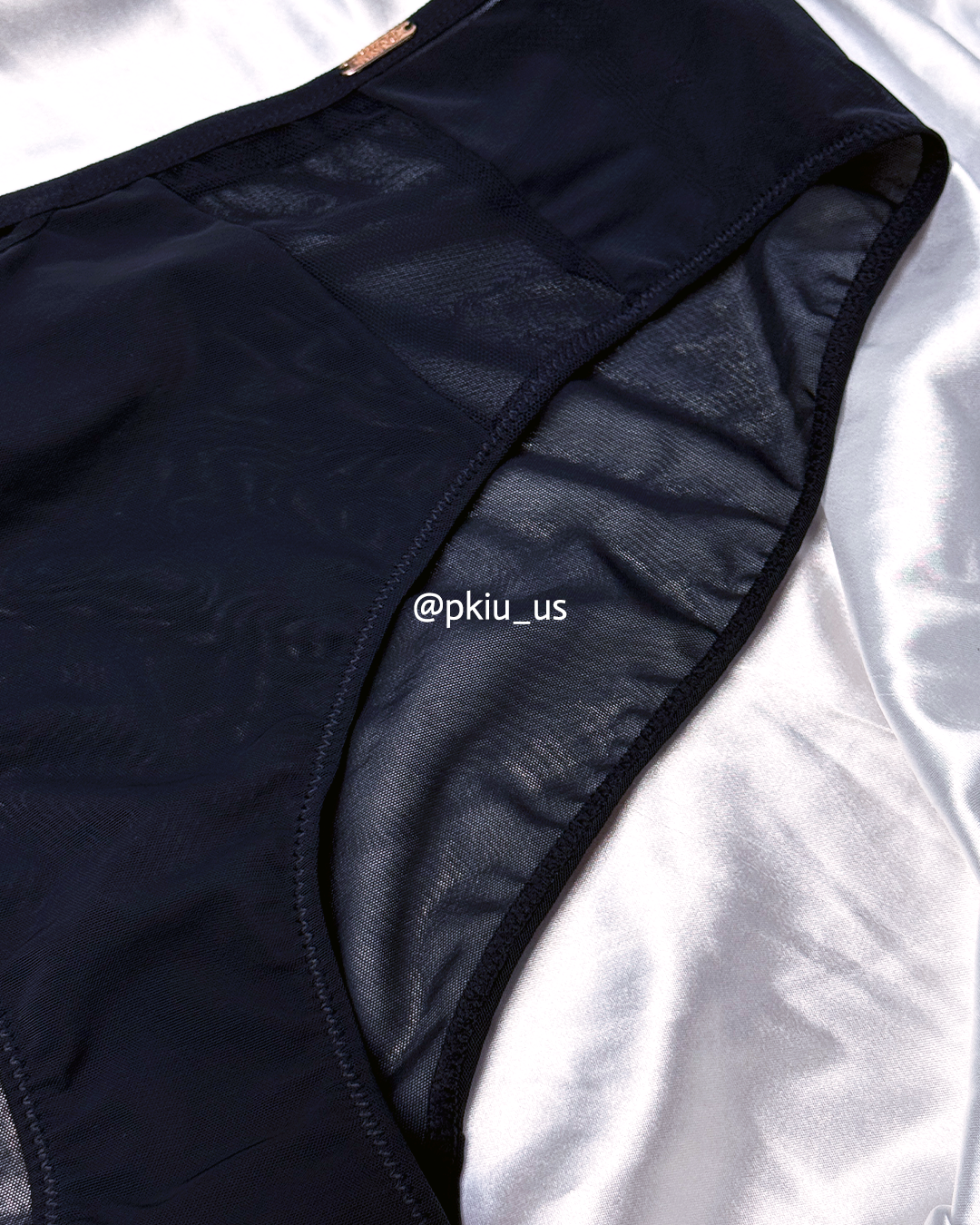 Close-up of a black high-waisted bikini bottom laid flat on a white satin fabric background, showing the sheer mesh side panel and black trim.