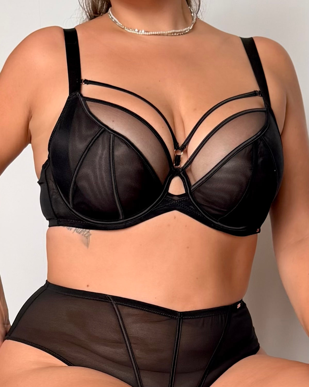 A close-up of a model wearing a black sheer mesh lingerie set with structured cups and strappy details, paired with matching high-waisted briefs.