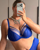 A woman taking a mirror selfie wearing a royal blue sheer mesh lingerie set with structured cups and a silver beaded fringe necklace.