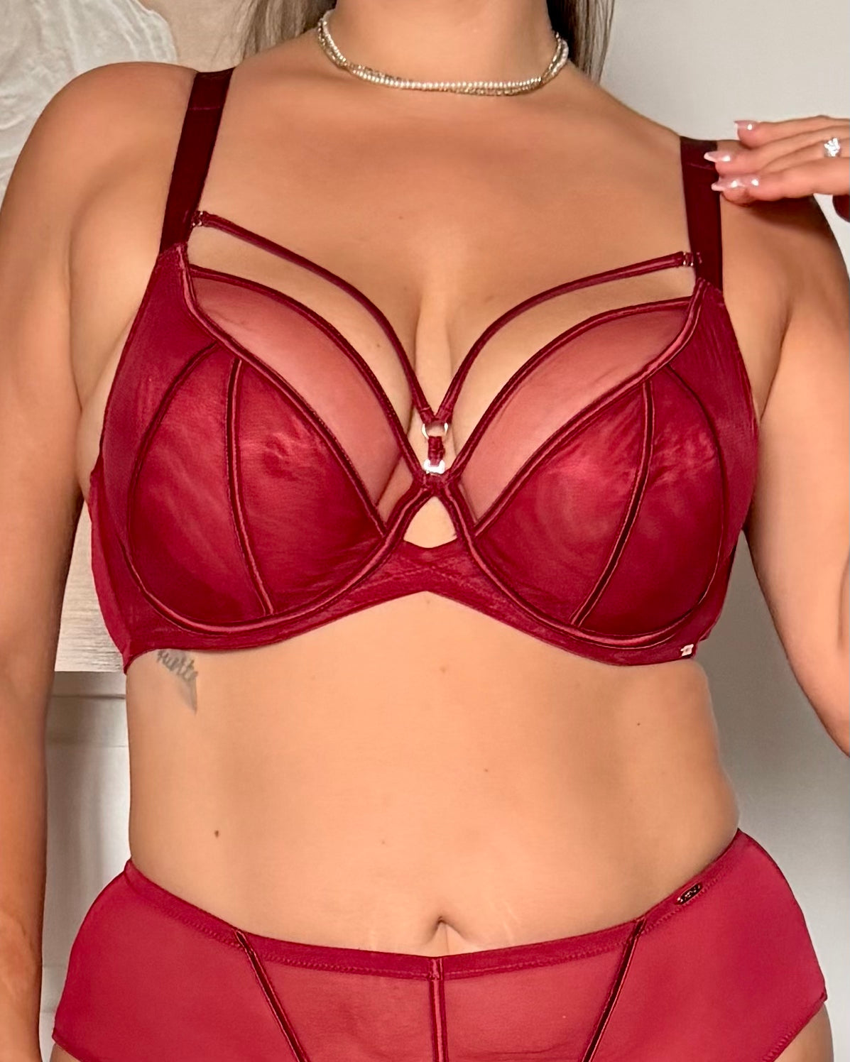 A close-up of a model wearing a sheer burgundy red strappy lingerie bra and matching underwear, featuring a small rhinestone center charm.