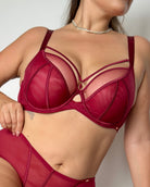 A close-up of a model wearing a structured burgundy lingerie set with sheer mesh panels and strappy details, paired with a matching high-waisted brief.